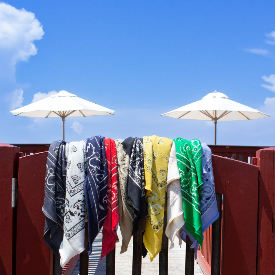 A row of colorful bandana towels hanging over a red wooden fence on a sunny beach deck, with white umbrellas and a blue sky in the background.