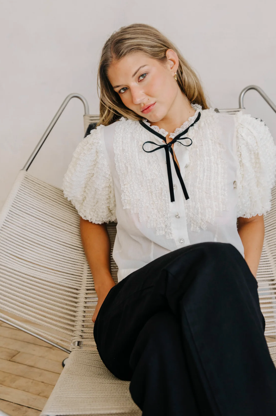Woman wearing a white blouse with black bow and black pants sitting on a chair. love the label ruffle white blouse