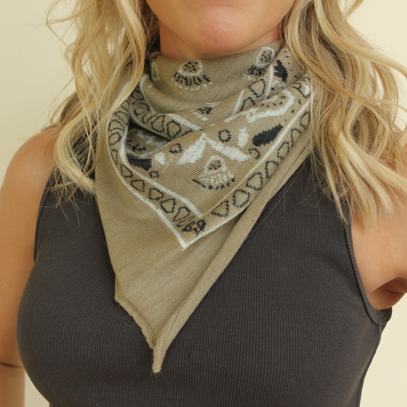 A close-up of a woman wearing a beige bandana-style neck gaiter with a black and white paisley pattern, paired with a dark gray ribbed tank top.