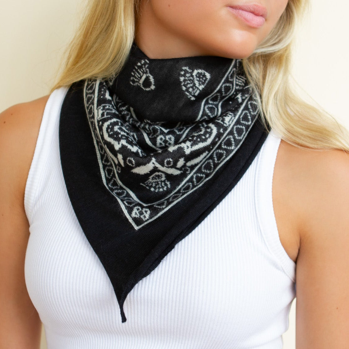 A blonde woman wearing a black bandana with a white paisley pattern around her neck, paired with a white ribbed tank top, set against a plain beige background.