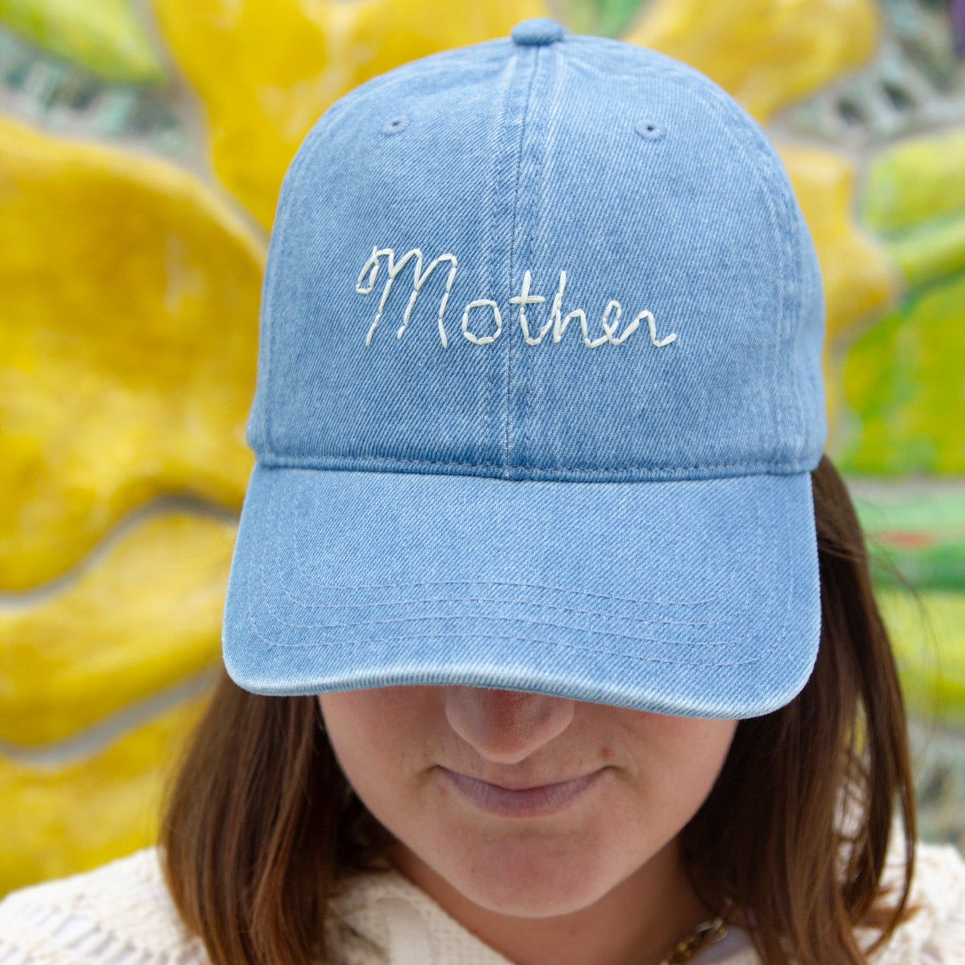 Light blue cap with 'Mother' embroidery worn by a person against a colorful background