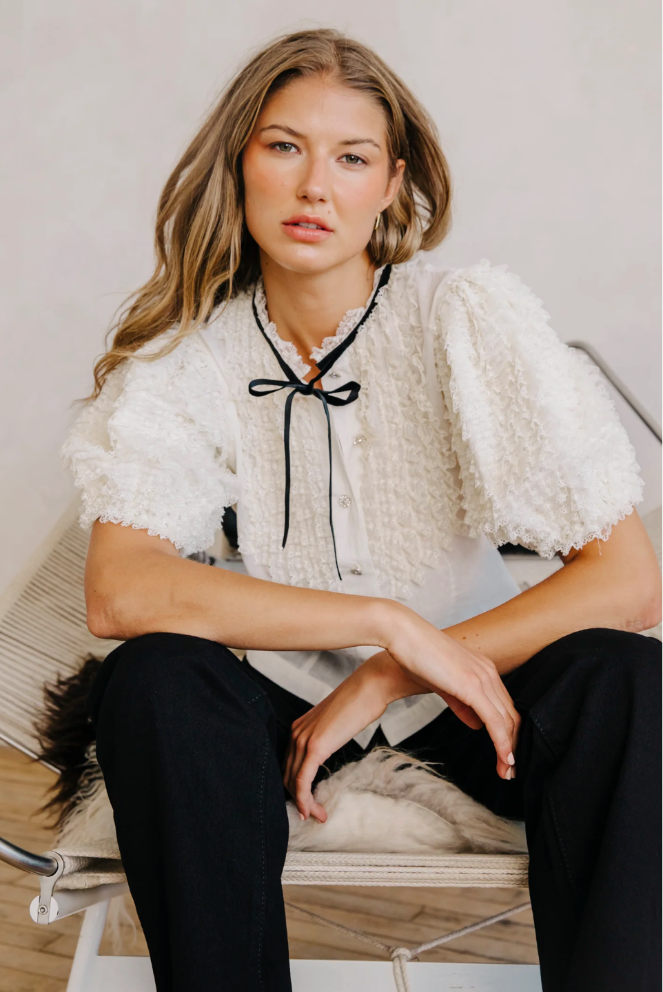 Woman wearing a white textured blouse with black trim and black pants sitting on a wooden chair. Love the label